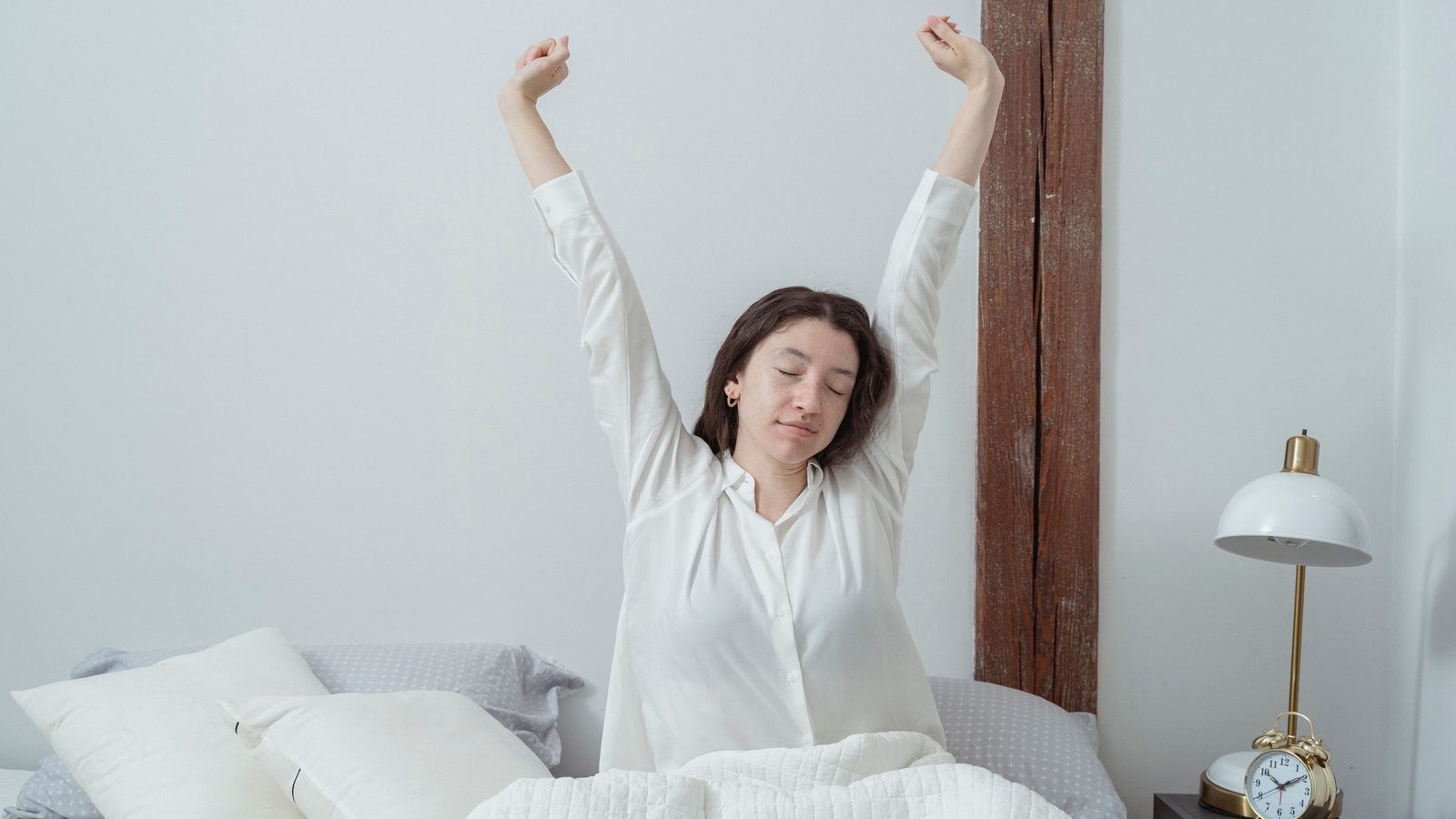 A woman stretching her arms overhead while sitting up in bed, with an alarm clock on the nightstand beside her