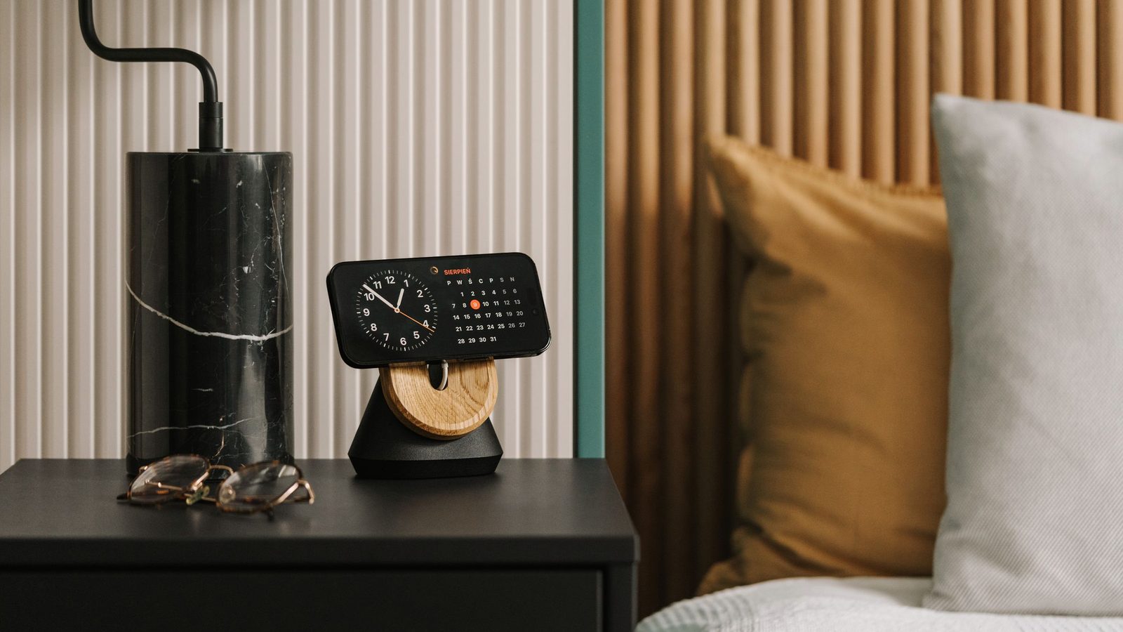 iPhone in MagSafe stand on bedside table displaying clock and calendar app, with bed pillows visible to the right