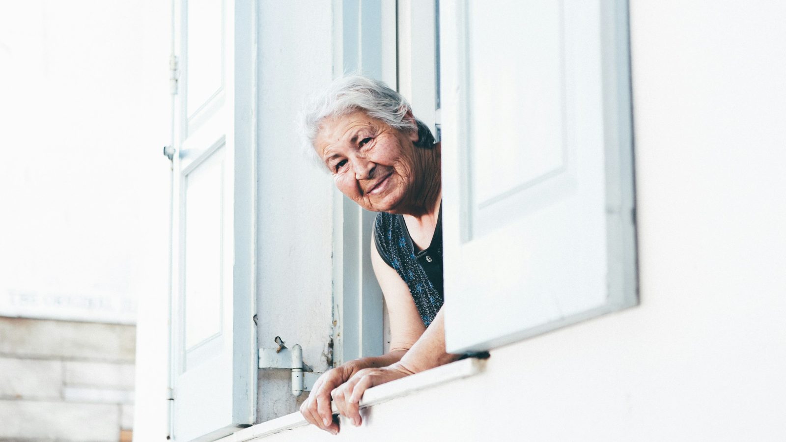 Older woman with white hair leaning out of a white window frame, smiling, with bright daylight behind her