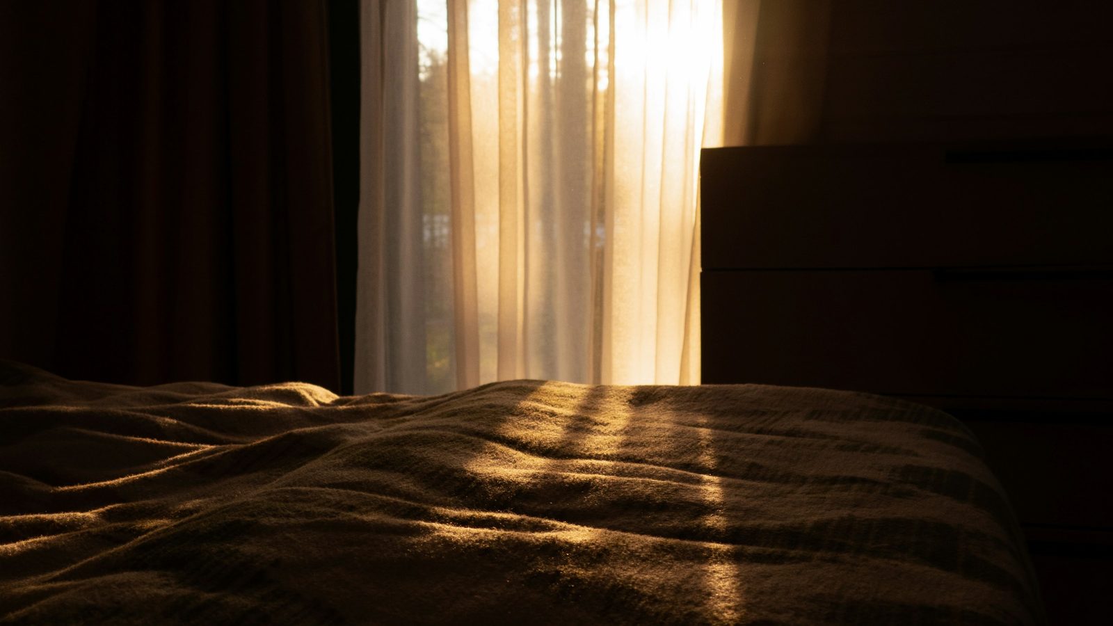 Soft morning light passing through sheer curtains onto an unmade bed