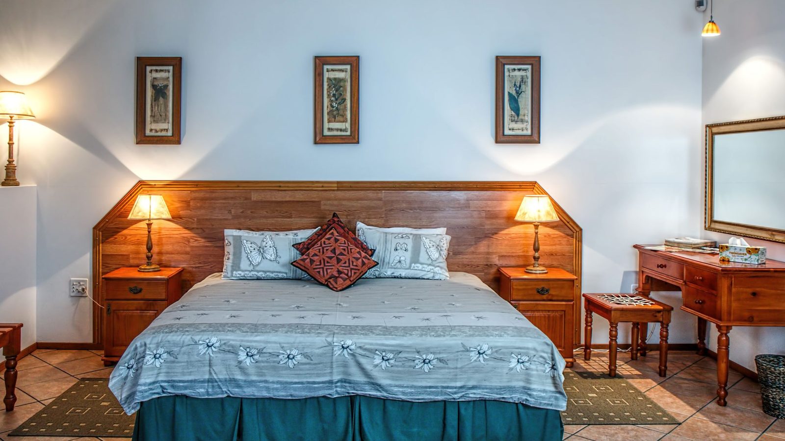 A neatly made bed in a bright bedroom with light blue bedding, decorative pillows, wooden nightstands with lamps on either side, and framed artwork on the walls