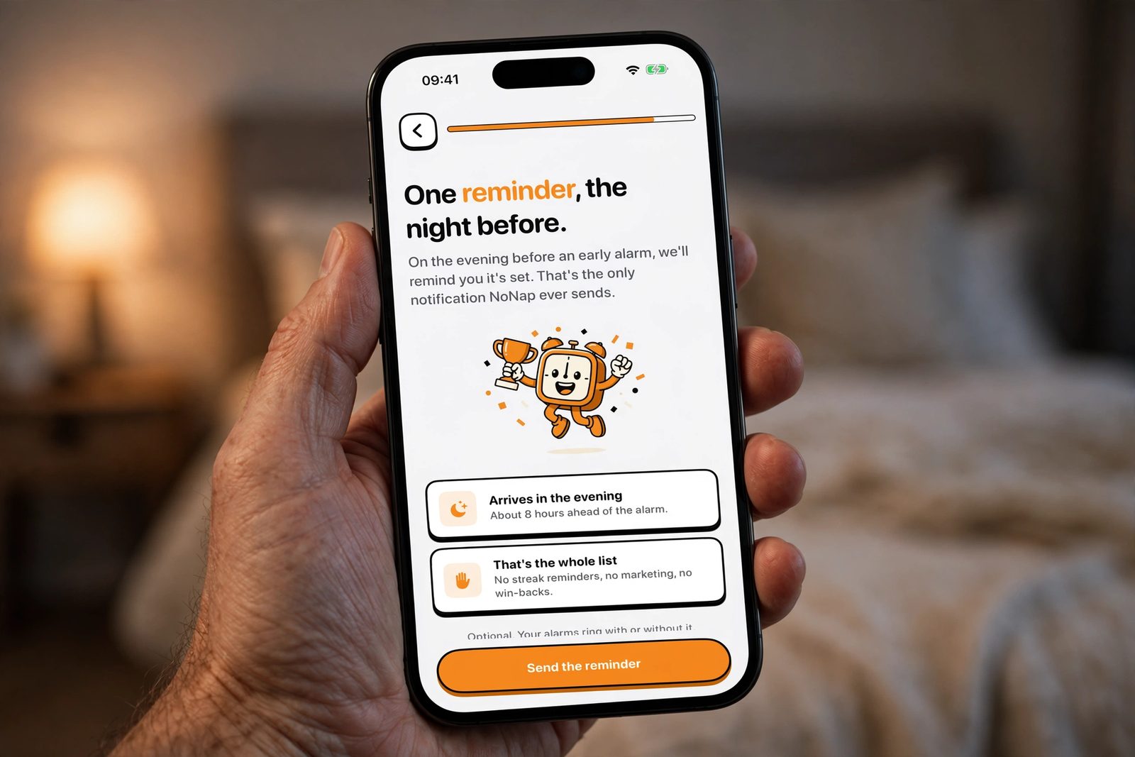 A hand holding an iPhone in a lamp-lit bedroom, showing NoNap's night-before reminder screen, sent about eight hours ahead of the alarm